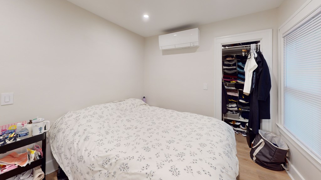A bedroom with a bed, a window with blinds, a coat rack with clothes, and a bag on the floor.