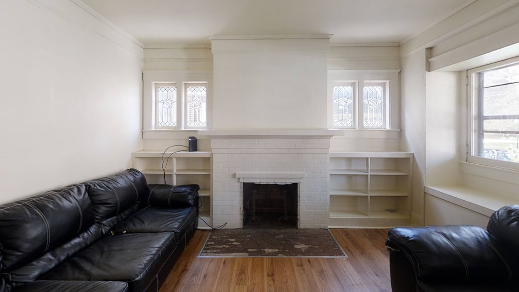 A black leather couch sits in front of a white fireplace in a room with wooden floors and white walls.