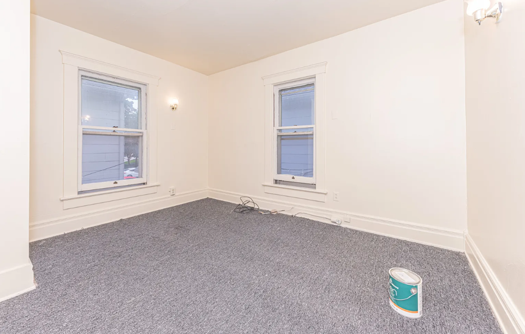 A room with a grey carpet and two windows.