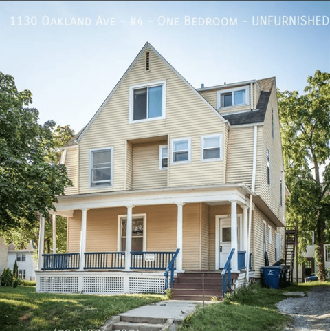 A two-story house with a porch and a blue trash can in front.