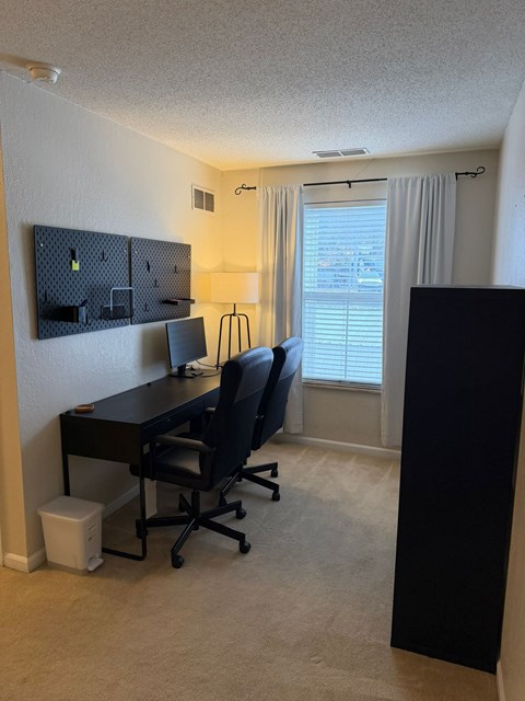A black desk with a black chair in a room with a white wall and a window with white blinds.
