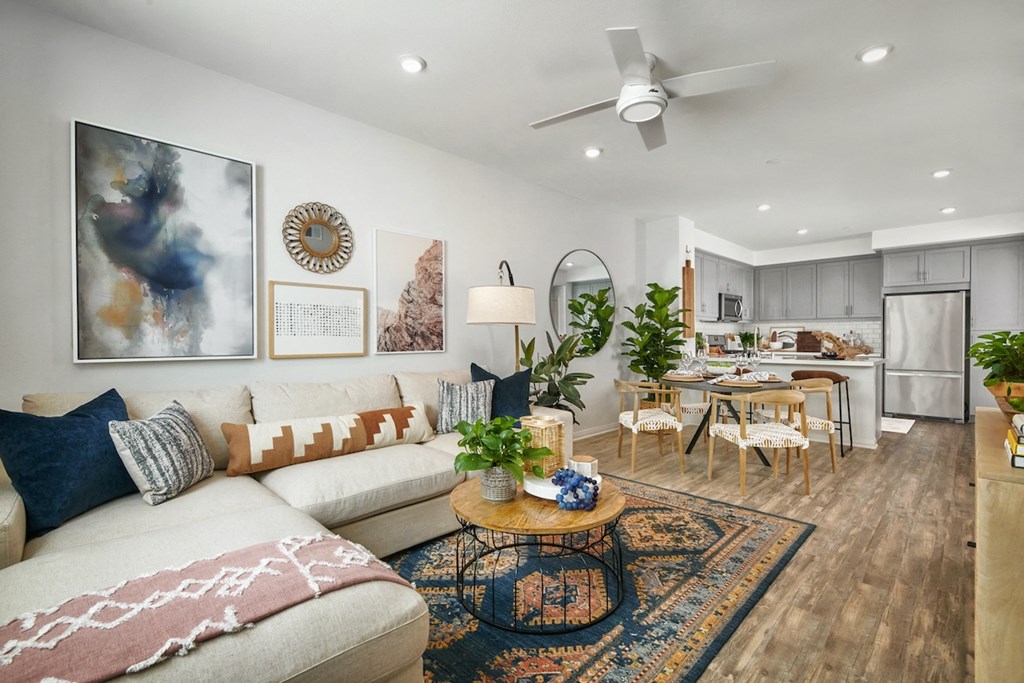 2 BR Apartments in Riverside CA - Lincoln Village - Furnished Living Room with White L-Shaped Couch, Round Wood-Topped Coffee Table, Oriental Rug, White Walls, Ceiling Fan, Dining Table with Chairs, Mounted Circular Mirror, House Plants, and View of Kitchen