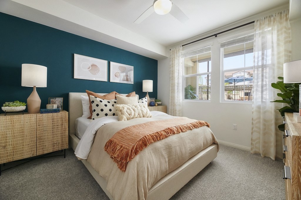 1 BR Apartments in Riverside CA - Lincoln Village - Furnished Bedroom with Carpeted Flooring, Large Bed, teal Accent Wall, Large Window, Wooden Dresser, Crocodile-Skinned Nightstands with Storage Cabinets, Lamps, and House Plant