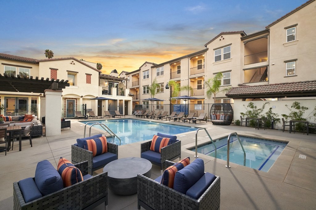 Apartments in Riverside, CA for Rent - Lincoln Village - Large Pool and Hot Tub, with Lounge Chairs, Outdoor Seating Area with Coffee Table, Covered Sofas, and Umbrellas, with White Building Exterior in the Background