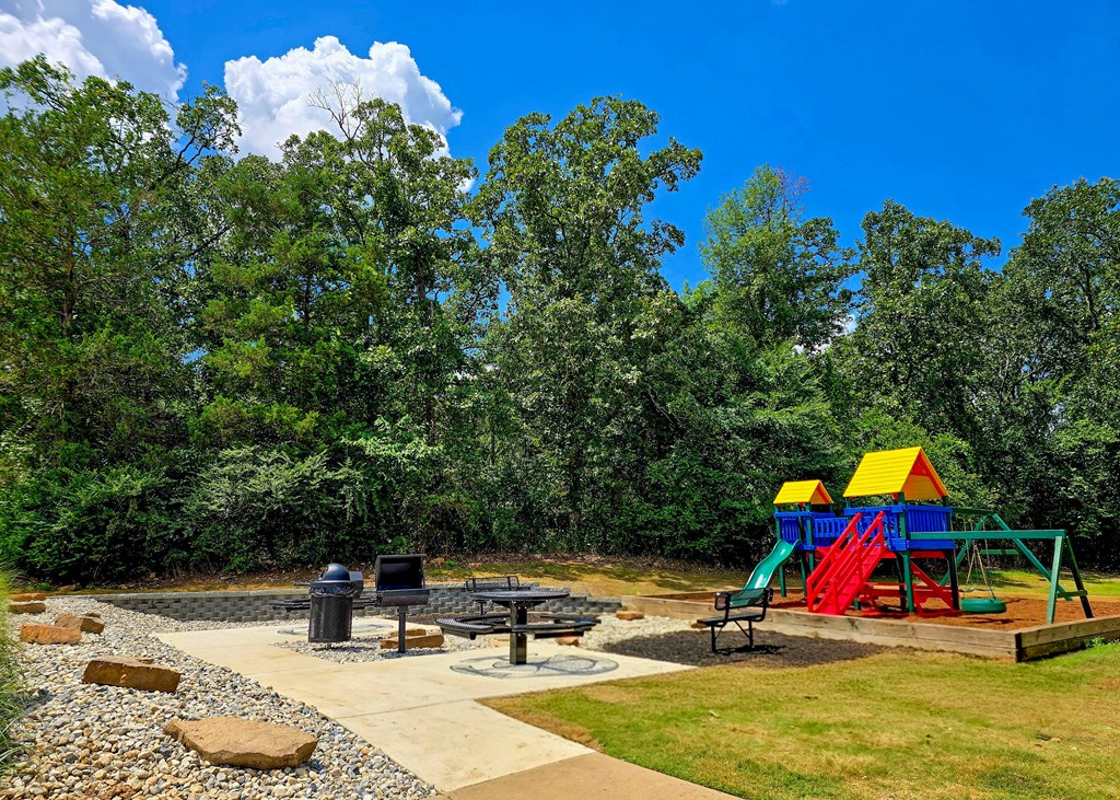 A playground with a slide, swings, and a picnic table is surrounded by trees.