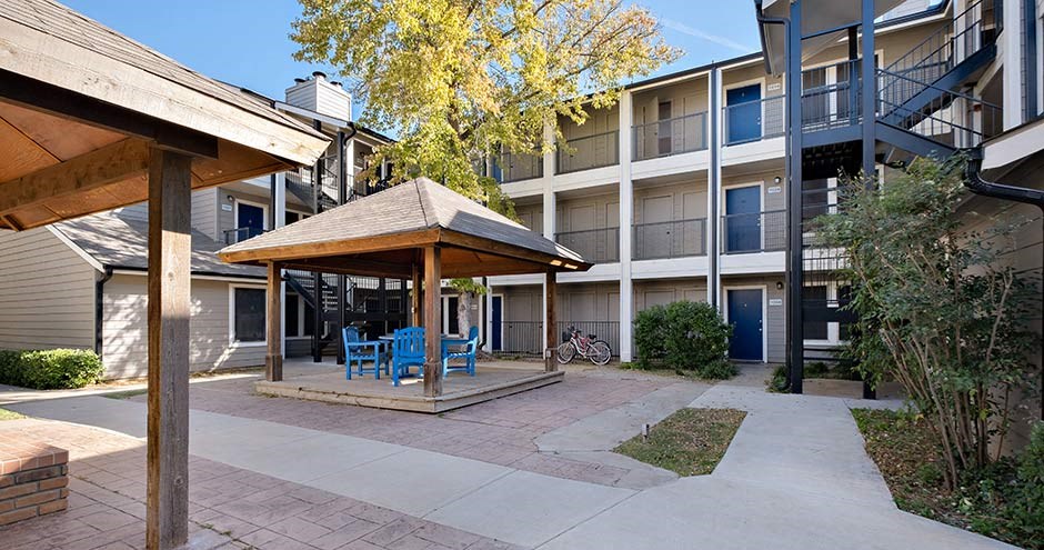 Cement red and beige courtyard with a wooden enclosed outdoor seating and bbq area against a sky blue background with green and yellow leaves on a tall tree