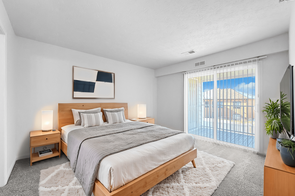 A bedroom with a large bed, a nightstand with a lamp, and a window with blinds.