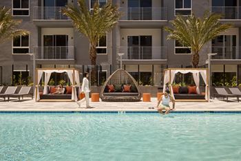 A woman is sitting on the edge of a pool in front of a building with palm trees.