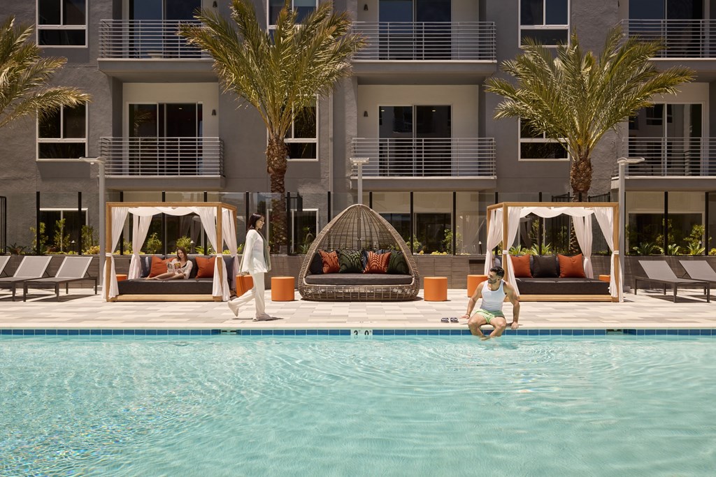A woman is sitting on the edge of a pool in front of a building with palm trees.