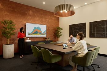 A woman in a red top stands in front of a whiteboard with a graph on it, while two other people sit at a table with laptops.