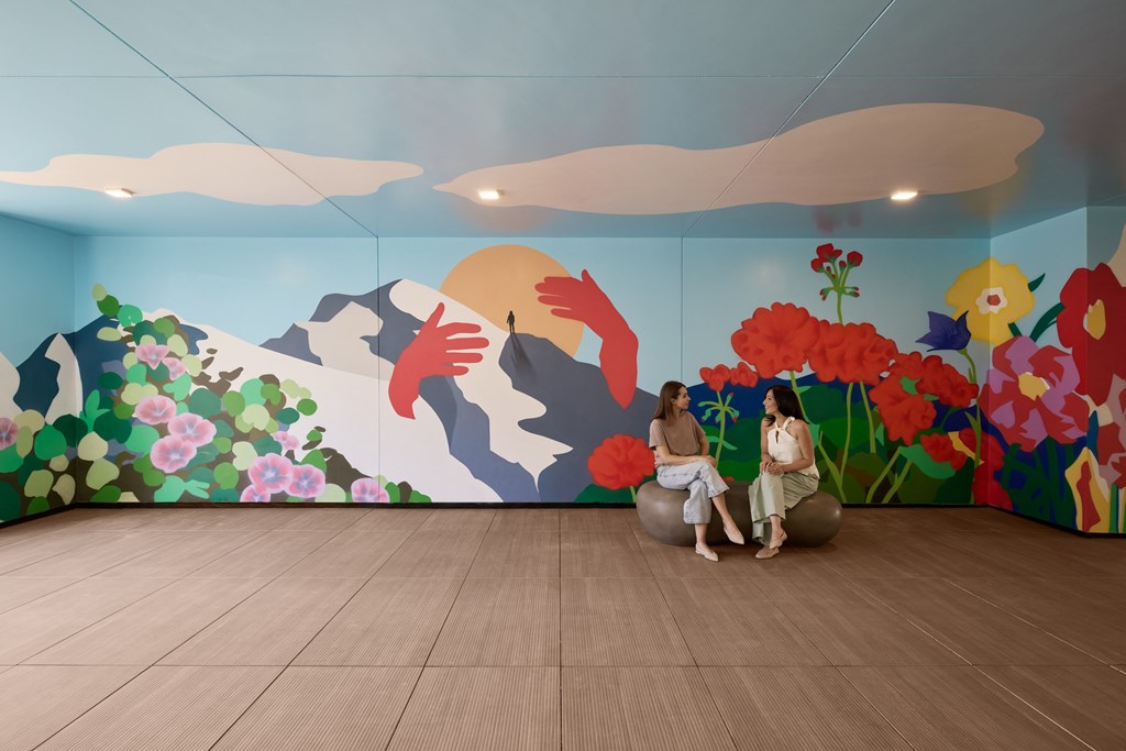 Two people sitting on a rock in a room with a painted mountain and flowers.