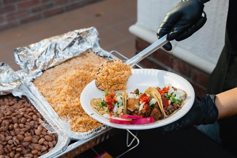 A person is serving food on a plate with a tray of beans and rice in the background.