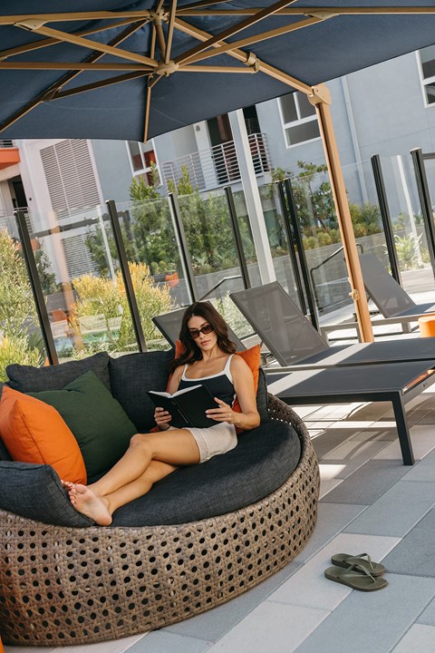A woman is sitting on a circular couch under a blue umbrella, reading a book.