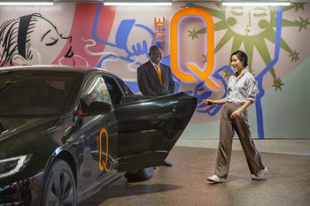 A woman is standing next to a black car with the letter Q on it.
