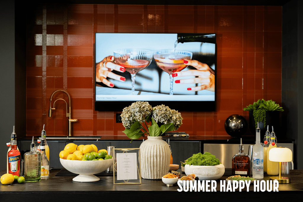 A kitchen counter with a bowl of lemons, a vase with flowers, and a bottle of alcohol with a TV screen showing a cocktail in the background.