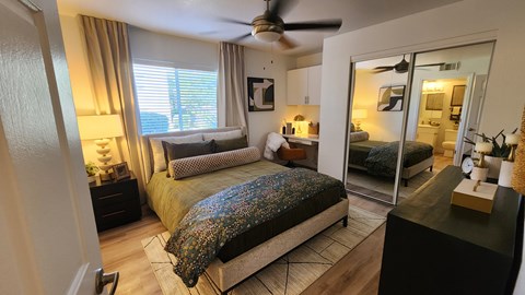 A bedroom with a bed covered in a multicolored blanket, a ceiling fan, and a mirrored doorway leading to a living area.