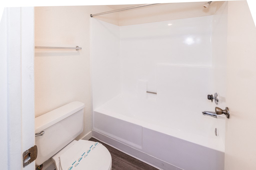 A white toilet with a blue line on the lid is in a small bathroom with a white bathtub and a white shower curtain.