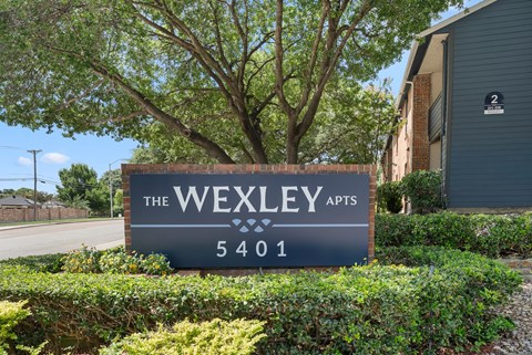 A sign for The Wexley Apts is in front of a tree and a building.