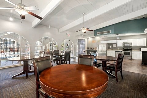 A large wooden table is surrounded by chairs in a spacious room with a kitchen in the background.