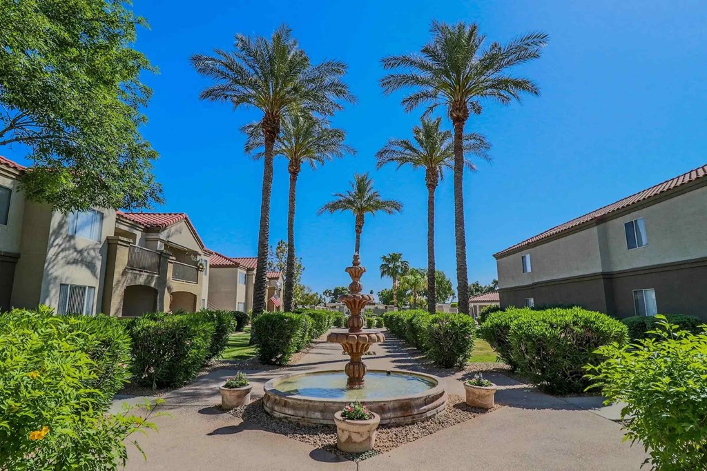 A fountain in the middle of a garden with palm trees and houses in the background.