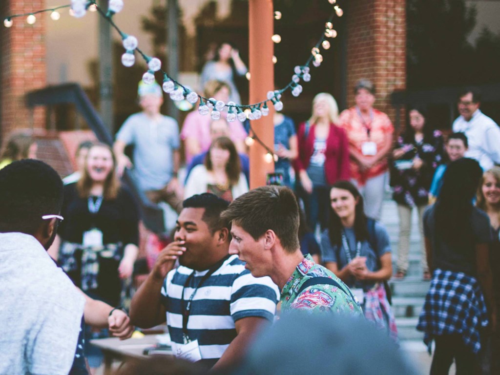 A large gathering of young people enjoys food and drinks at outdoor tables