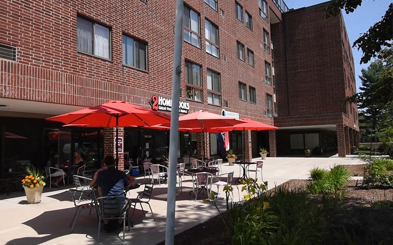 A man is sitting at a table under a red umbrella in front of a brick building.