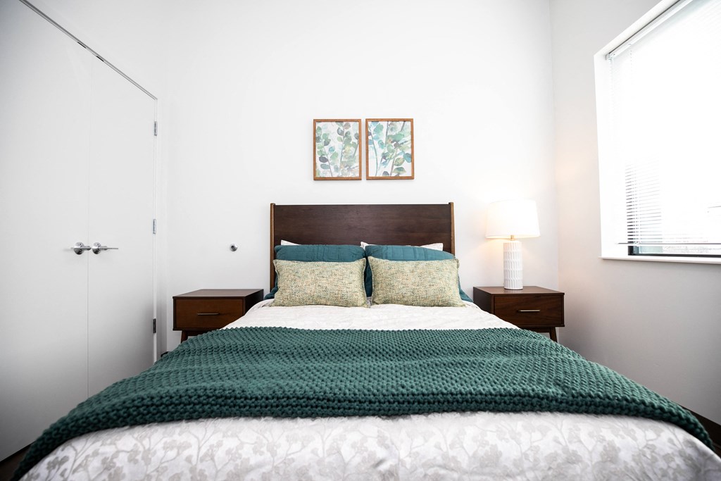 A bed with a green blanket and two pillows sits in a room with white walls.