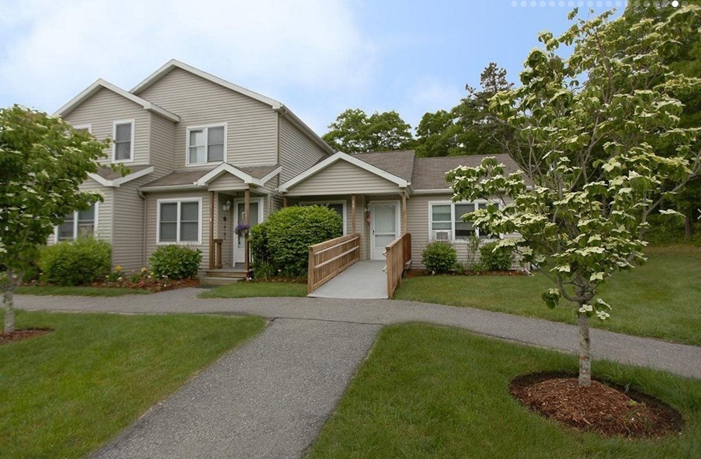 A house with a grey roof and a white door is surrounded by greenery.