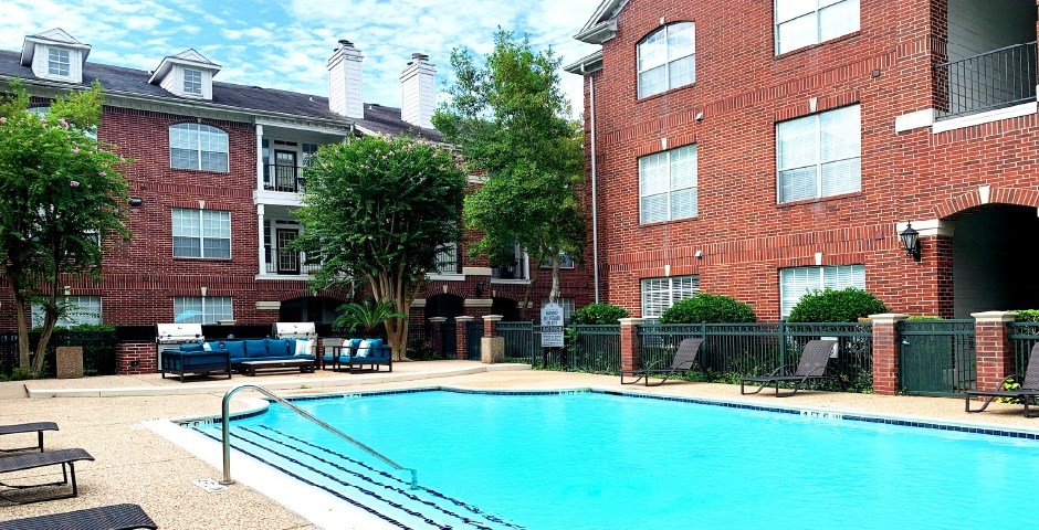 Luxury apartments with red brick exteriors, large swimming pool with spacious sundeck, grill, beautiful landscaping, lush landscaping, chaste trees, and pool-side lounge seating at Tuscany Apartments in Houston.