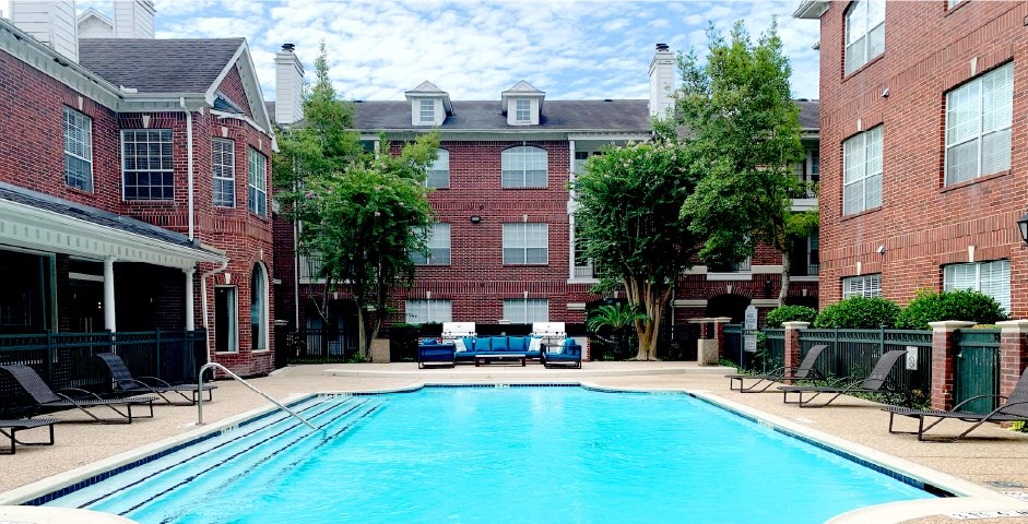 Luxury apartments with red brick exteriors, large swimming pool with spacious sundeck, grill, beautiful landscaping, lush landscaping, chaste trees, and pool-side lounge seating at Tuscany Apartments in Houston.