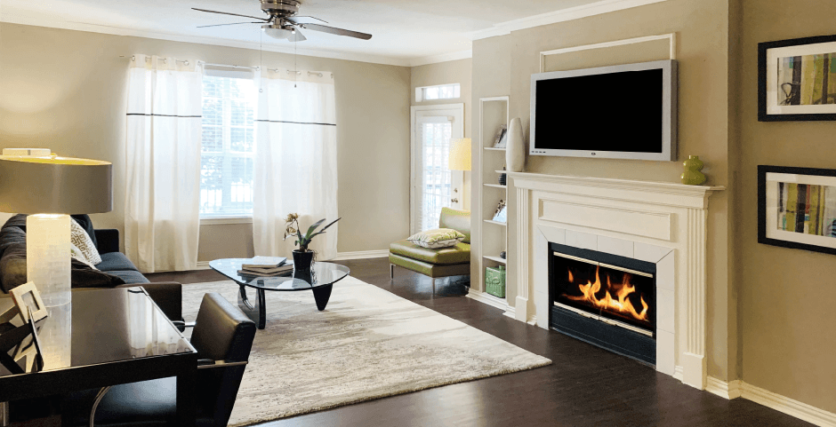 Spacious living room with wood-burning fireplace with glass doors, 9-foot ceilings, built-in shelves, and wood grain plank flooring at The Lanesborough Apartments in Houston.
