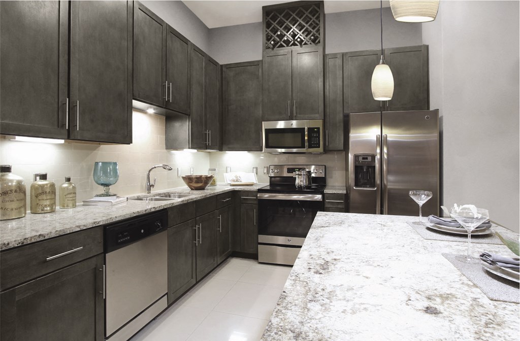 An open plan kitchen with white granite countertops, overhead and under the counter dark wood cabinetry, steel appliances, and built-in wineracks