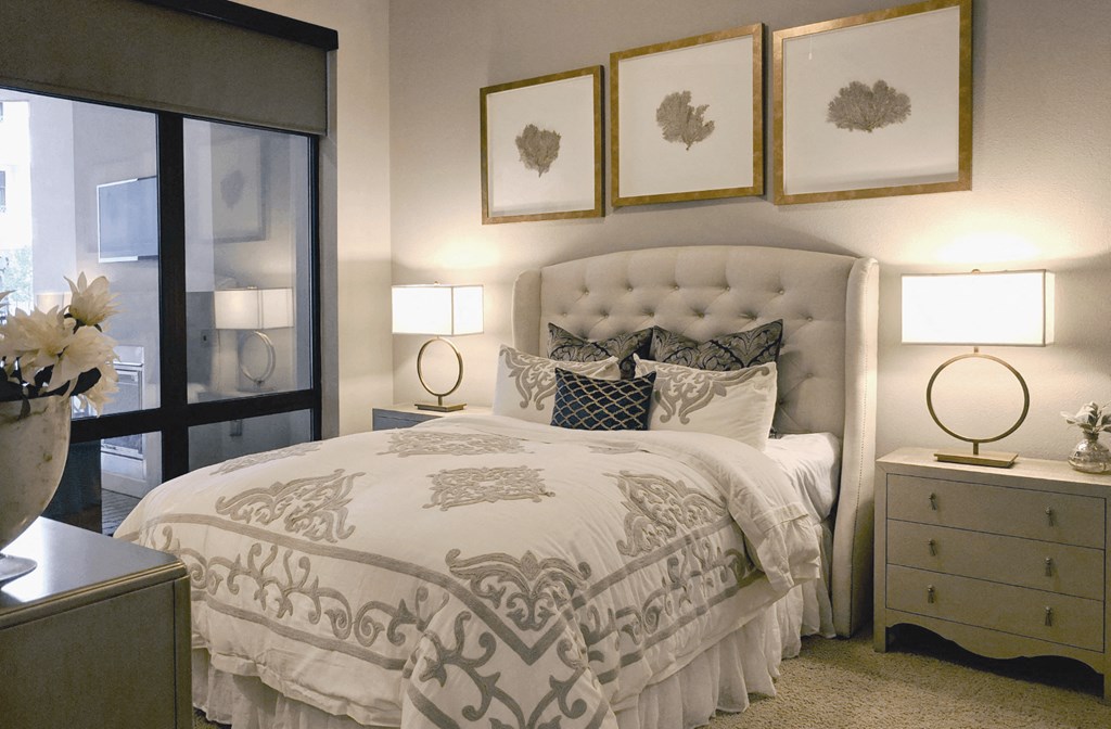 A carpeted bedroom with a king-size plush bed and a dresser overlooking a patio through a glass floor to ceiling window