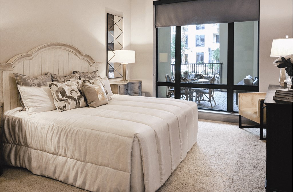 A carpeted bedroom with a king-size plush bed and a dresser overlooking a patio through a glass floor to ceiling window