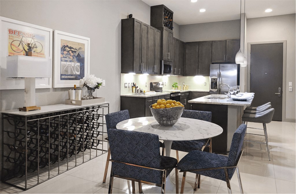 A round dining table with four chairs, a long wine racks along a wall and a view of an open kitchen with dark wood cabinetry