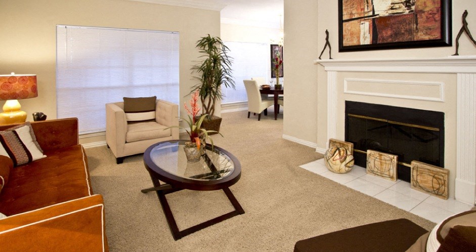 Spacious living room with wood-burning fireplace with glass doors, ceiling fan, and 9-foot ceiling at Tuscany Lane Apartments in Houston.