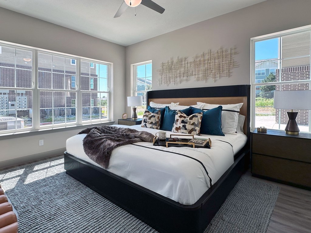 A bedroom with a large bed, a ceiling fan, and a view of a cityscape through the windows.