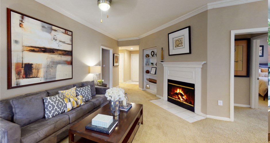 Spacious living room with wood-burning fireplace with glass doors, ceiling fan, crown molding, and built-in bookshelves at Tuscany Apartments in Houston.