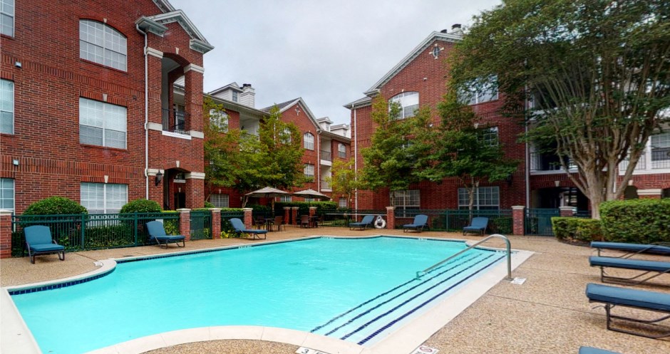 Luxury apartments with red brick exteriors, large swimming pool with spacious sundeck, grill, beautiful landscaping, lush landscaping, chaste trees, and pool-side lounge seating at Tuscany Apartments in Houston.