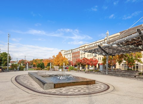 A fountain in the middle of a plaza with buildings and trees in the background.