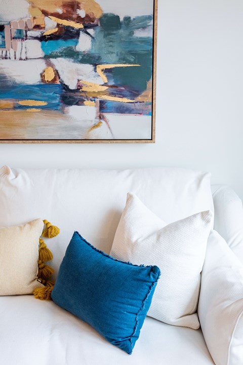 A painting hangs on a wall above a white couch with pillows.