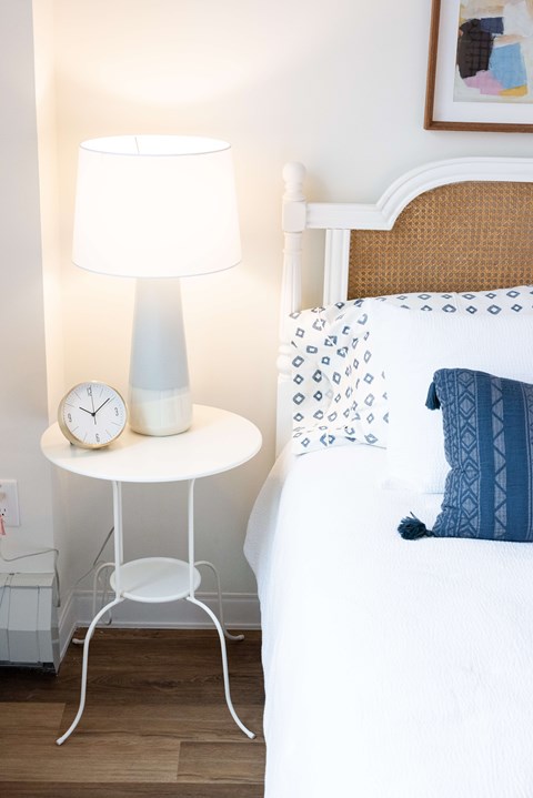 A white bedside table with a lamp and clock beside a bed.