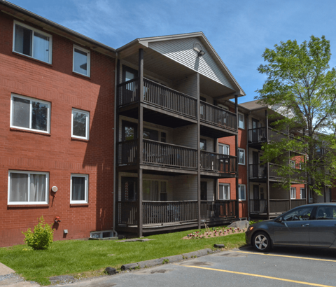 A grey car is parked in a parking lot in front of a red brick building with a balcony.