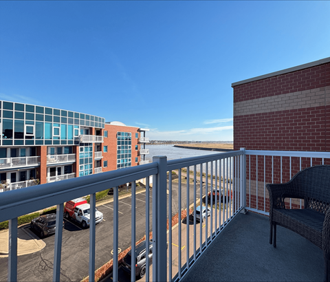 A balcony with a chair and a railing overlooking a parking lot and buildings.