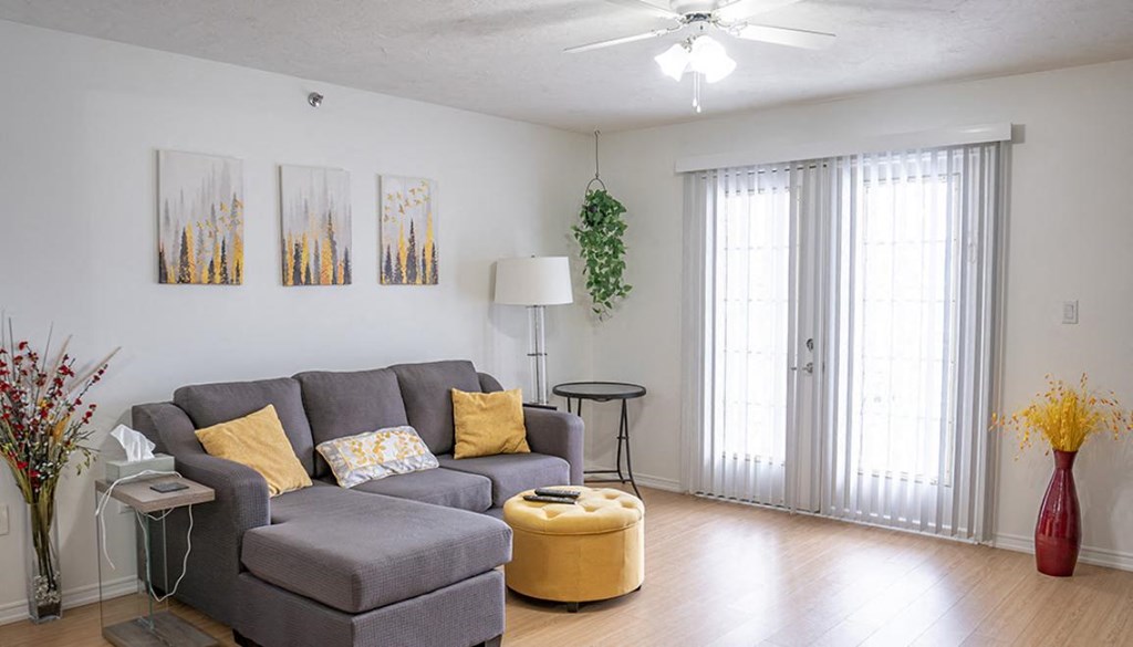 A living room with a grey couch, yellow ottoman, and a ceiling fan.