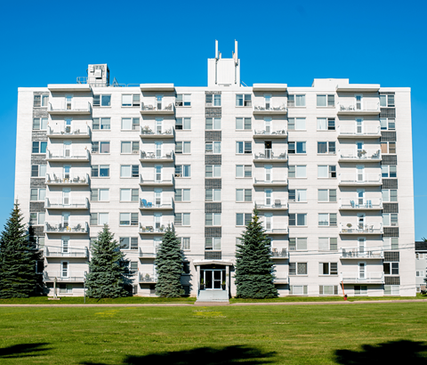 A large white apartment building with balconies and trees in front.