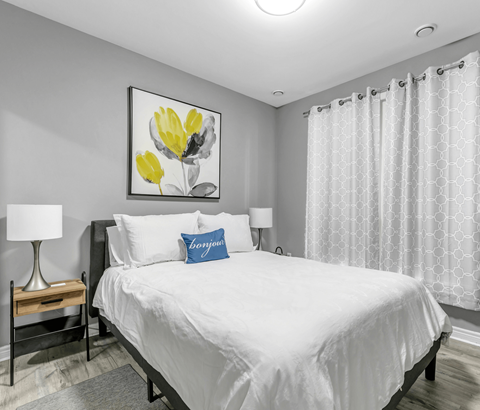 A bedroom with a large bed, a nightstand with a lamp, and a framed picture of yellow flowers on the wall.