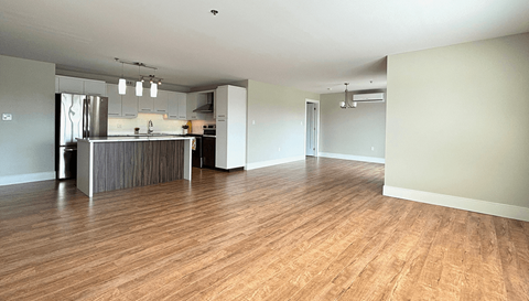 A spacious room with wooden flooring and a kitchen area in the background.