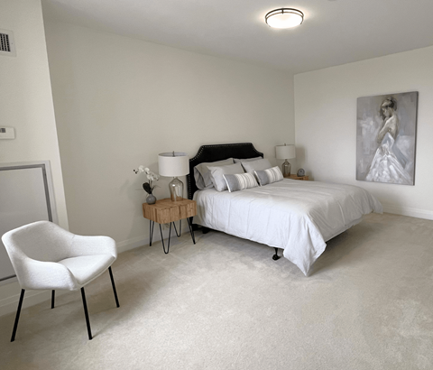 A white chair sits in a bedroom with a bed and a painting on the wall.