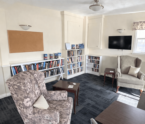 A living room with a grey floral armchair, a wooden coffee table, a grey sofa, a bookshelf with books, a television, and a window with curtains.